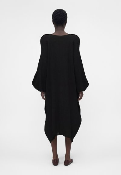 Άτομο που στέκεται με την πλάτη γυρισμένη προς τα εμπρός, φοράει μακρύ μαύρο φόρεμα με φουσκωτά μανίκια και σκοτεινά επίπεδα παπούτσια σε απλό λευκό φόντο.