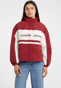 Rød og hvid quilted jakke med høj krave, med "Guess Jeans" logo og elastiske manchetter, parret med blå denim jeans.