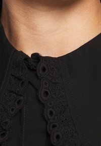 Tissu noir avec une bordure en dentelle brodée texturée le long de l'encolure. Petite fermeture par bouton au centre, drapé doux.