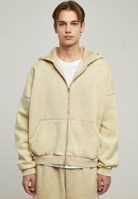 Beige zip-up hoodie with a spacious fit, soft texture, large hood, and kangaroo pockets, paired with a white t-shirt underneath.