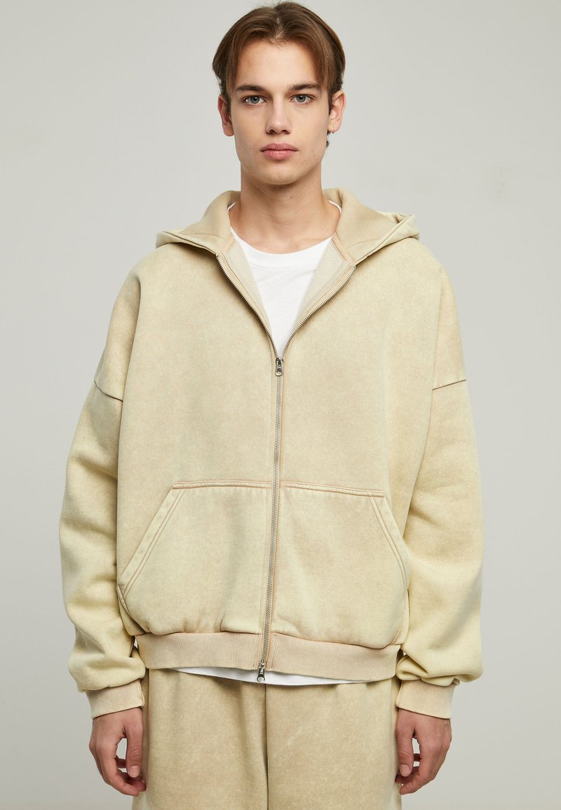 Sudadera con cremallera color beige de corte amplio, textura suave, gran capucha y bolsillos tipo canguro, combinada con una camiseta blanca debajo.