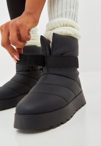 Botas de puff negro con un acabado suave y mate, que cuentan con una amplia correa de Velcro y una suela de goma estriada. Los acentos de costura añaden textura.