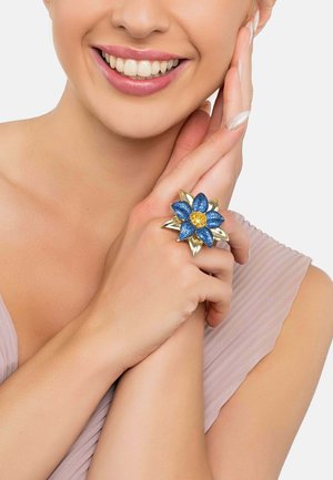 Blue and gold flower ring with textured blue petals and a central gold accent, set on a metallic base, worn on a hand.