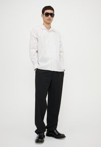 Camisa blanca a rayas con botones, pantalones anchos negros y zapatos negros brillantes. El modelo lleva gafas de sol negras. Diseño minimalista y colores neutros.