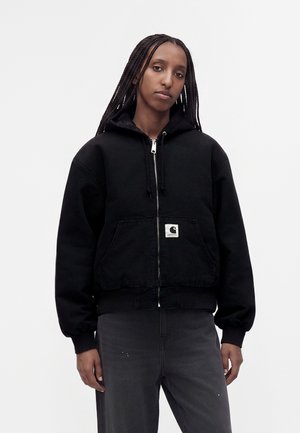 Personne avec de longues tresses portant un sweat à capuche noir zippé avec une poche avant et un pantalon gris foncé sur un fond blanc uni.