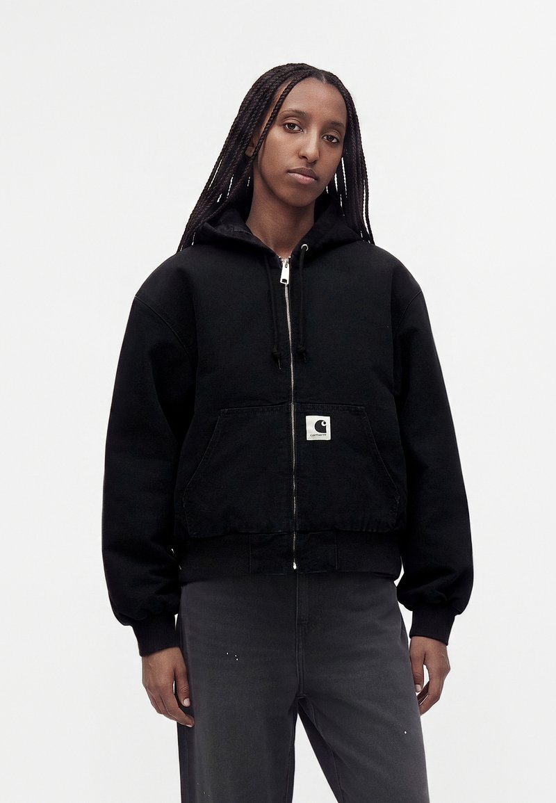 Personne avec de longues tresses portant un sweat à capuche noir zippé avec une poche avant et un pantalon gris foncé sur un fond blanc uni.