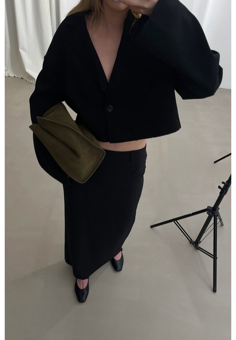 Personne portant une veste noire courte, une jupe noire taille haute et des chaussures noires, tenant une grande pochette vert olive à l’intérieur.