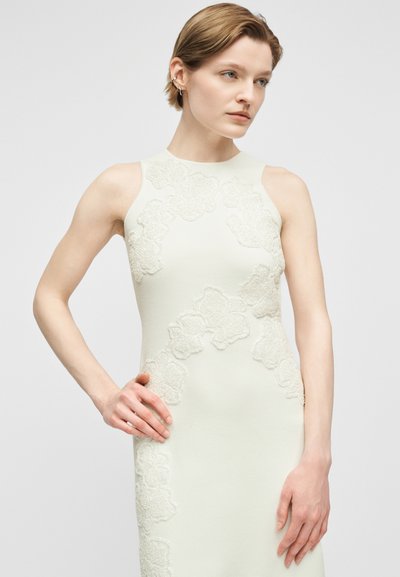 Νεαρή γυναίκα με κοντά μαλλιά, φορώντας άσπρο αμάνικο φόρεμα με λουλουδένια δαντελένια σχέδια, στέκεται μπροστά σε απλό φωτεινό γκρι φόντο.