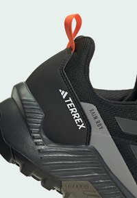 Sort atletisk sko med en struktureret overdel, grå detaljer og en lys orange trækstrop. Inkluderer branding "TERREX" og "RAIN.RDY."