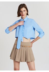 Camicetta azzurro chiaro tagliata con una piccola tasca, abbinata a una gonna plissettata beige con una cintura color muschio. Tessuto morbido e design su misura.