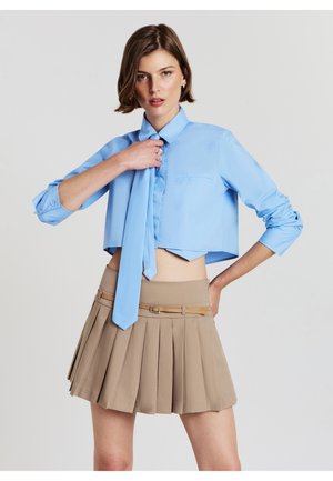 Camicetta azzurro chiaro tagliata con una piccola tasca, abbinata a una gonna plissettata beige con una cintura color muschio. Tessuto morbido e design su misura.