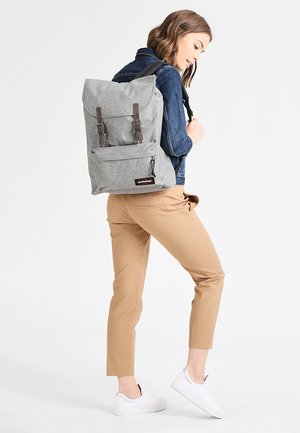 Gray fabric backpack with a roll-top design, featuring brown leather straps, front pocket, and a logo patch. Worn by a person.