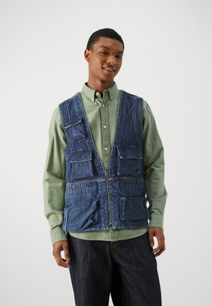 Jeansweste mit mehreren Taschen, versehen mit einem Reißverschluss, getragen über einem hellgrünen Hemd. Der Stoff hat ein leicht gebleichtes Aussehen.