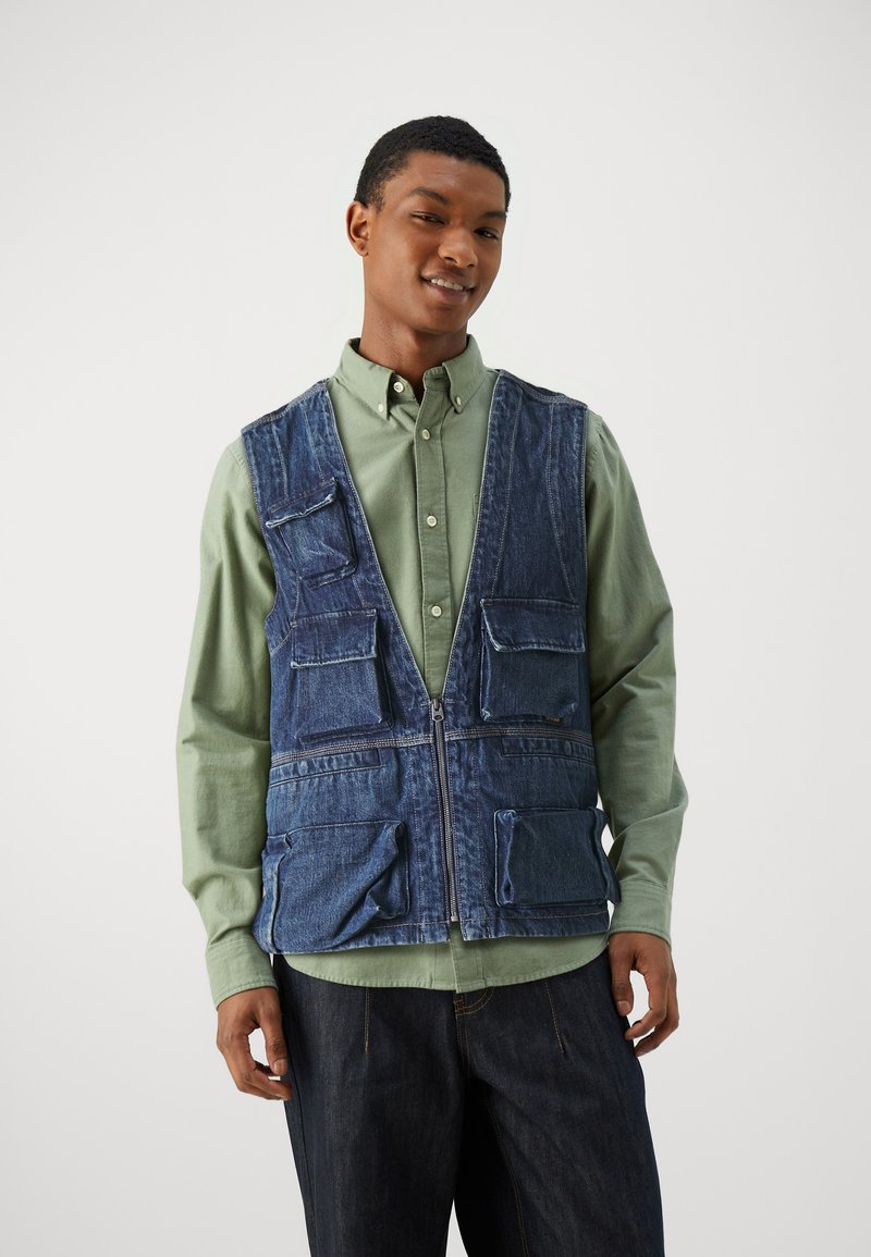 Denimvest med flere lommer, med lynlås lukning, båret over en lys grøn skjorte. Stoffet har et let falmet udseende.