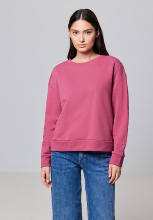 Sweatshirt rose avec clous sur les épaules, col rond et ourlet côtelé. Associé à un jean bleu à jambes larges ; le mannequin a les cheveux droits et foncés.