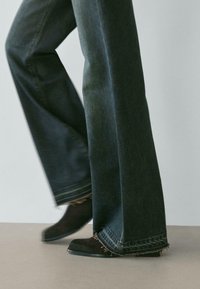 Osoba nosząca ciemne dzwoniaste dżinsy z postrzępionymi brzegami i czarne zamszowe buty stojąca na neutralnej podłodze na jednolitym tle.