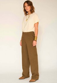 Femme portant un haut crème décontracté et un pantalon marron à jambes larges, présentant une texture lisse. Les accessoires incluent des colliers et bracelets en or superposés.