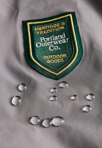 Sivo vodoodpudivé tkaniny s vyšitým zeleno-zlatým logem ve tvaru štítu s nápisem "Portland Outerwear Co. OUTDOOR GOODS." Viditelné kapky vody.