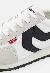 Leichte Sneaker mit einem weißen Obermaterial aus Stoff, einer grauen Wildleder-Spitze, schwarzen Lederakzenten und flachen weißen Schnürsenkeln. Rote Etikett-Details vorhanden.