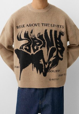 Persona vistiendo un suéter beige con un diseño abstracto negro y la frase "SUPÉRATE SOBRE LOS LÍMITES" en el pecho, combinado con jeans azules.