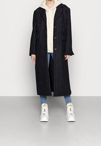 Lungo cappotto blu navy con strisce bianche, due tasche frontali, rever a punta e chiusura con bottone, indossato sopra una leggera felpa con cappuccio e jeans.