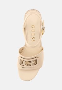 Sandalo con tacco beige, dotato di una larga fascia con logo dorato, design slingback, texture liscia e punta leggermente arrotondata.