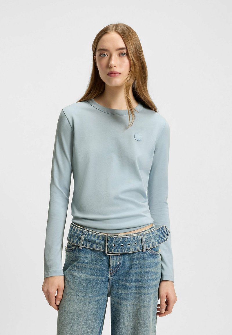 Helles blaues Langarmshirt aus weichem Stoff, mit rundem Halsausschnitt und einem kleinen runden Logo auf der Brust, kombiniert mit hoch geschnittenen Jeans.