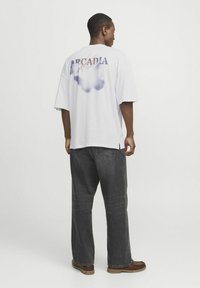 Camiseta de algodón blanca con corte oversize, con "ARCADIA" impreso en la parte posterior en azul, con un degradado desvanecido y diseño curvado.