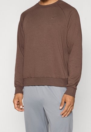 Hombre vistiendo un sudadero marrón de manga larga de Nike y pantalones gris claro, de pie con los brazos relajados a los lados.