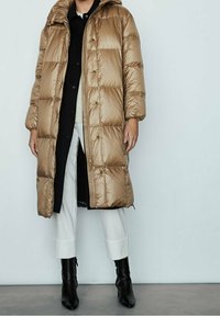 Cappotto lungo, imbottito, in tessuto lucido color tan con cappuccio, dotato di zip frontale, tasche laterali e design sotto al ginocchio, abbinato a stivali neri.