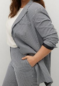 Kurtka w czarno-białą kratkę w stylu gingham z pasującymi spodniami. Zawiera kieszeń z przodu, klapy oraz mankiety z jednolitymi czarnymi akcentami.