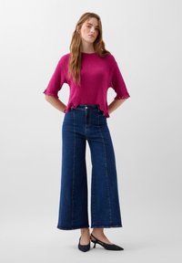 Maglione fucsia a maglia con maniche corte e bordi ondulati, abbinato a jeans scuri a vita alta e gamba ampia e scarpe slingback blu navy.