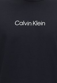Fekete szövet, fehér "Calvin Klein" logó szöveggel, amely az öltözék felső mellrészének közepén helyezkedik el.
