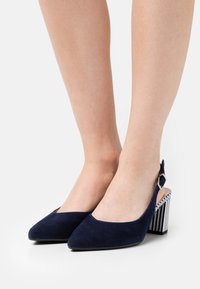 Tacchi slingback in suede blu navy con punta affusolata e tacco a strisce robusto. Presentano una fibbia laterale argentata per la chiusura.