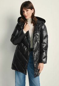 Czarna puchowa kurtka o błyszczącej powierzchni, z kapturem i wzorem w zygzaki. Noszona z jasnym golfem i niebieskimi dżinsami.