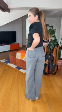 Femme aux longs cheveux attachés en queue de cheval, portant un haut noir et un pantalon gris à jambes larges, debout à l'intérieur avec les mains sur les hanches, près des plantes et des meubles.