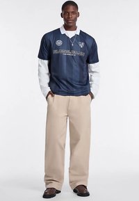 Marineblå sportstrøje med hvid krave og logo, kombineret med beige bukser med vide ben og mørke sko. Trøjen har vertikale striber og tekst.