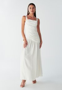 Maxi šaty v bílé barvě s nařaseným živůtkem, tenkými ramínky a splývavou sukní. Kombinovány se zlatými sandály na pásky.