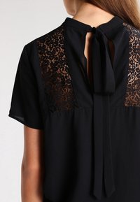 Blusa negra con detalles de encaje en la parte posterior y mangas cortas, con un cierre de lazo en el cuello y una textura de tela suave.