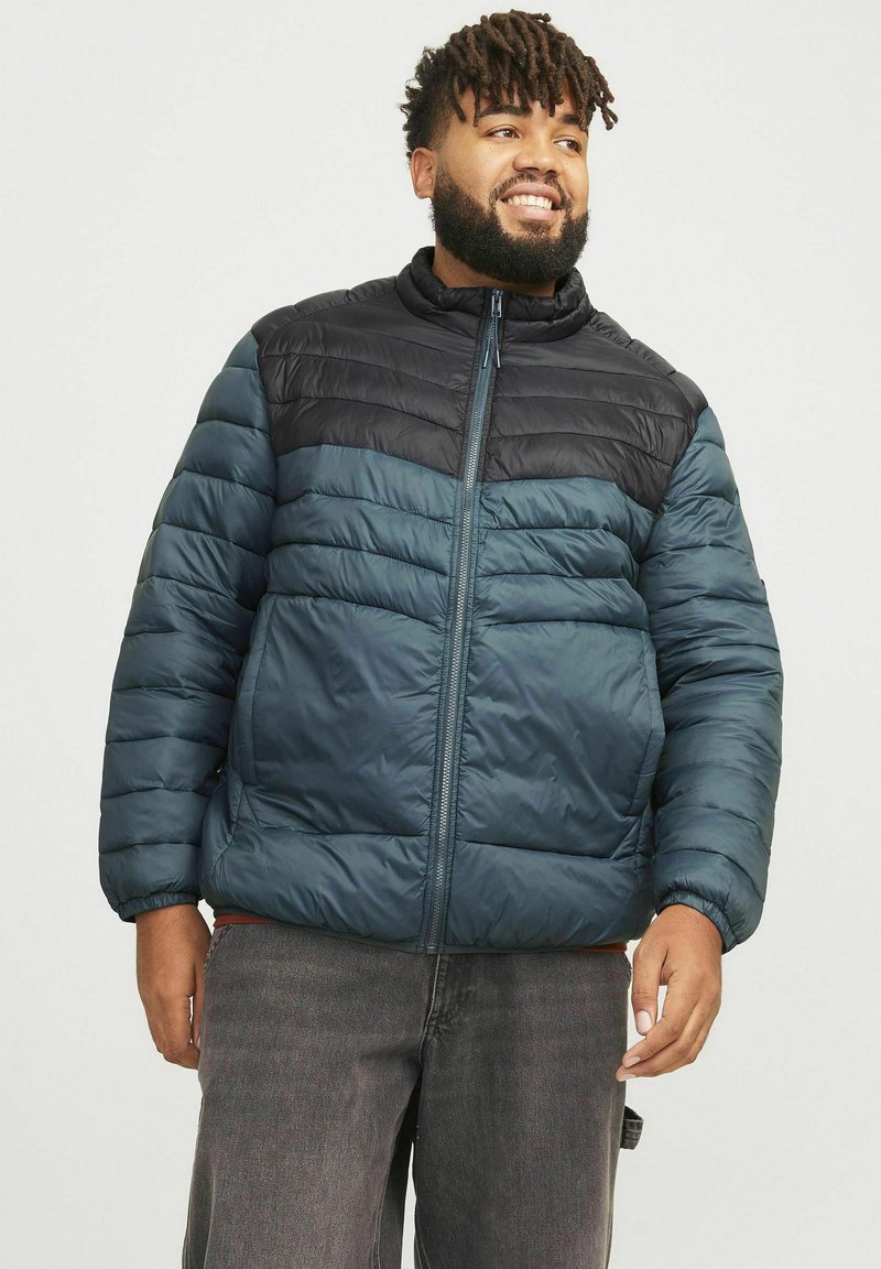 Puffjakke med mørkeblå og sorte paneler, høj krave, lynlåslukning og kviltet tekstur. Bæres over mørkegrå jeans, som viser varme og stil.