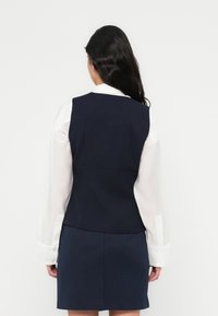 Σκούφο μπλε γιλέκο με ραβδωτή υφή, φορεμένο πάνω από λευκό πουκάμισο με γιακά. Διαθέτει εφαρμοστή σχεδίαση και ομαλό ύφασμα. Φούστα στο ίδιο μπλε χρώμα.