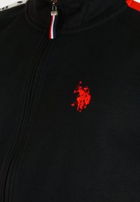 Giubbotto zip-up nero con logo ricamato rosso, caratterizzato da un colletto alto e accenti rossi a contrasto sulle spalle. Realizzato in tessuto morbido e texturizzato.