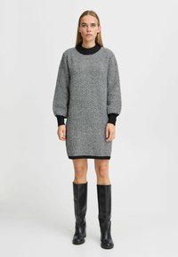 Schwarz-weißer Hahnentritt-Strickkleid mit langen Ballonärmeln und einem schwarzen, runden Kragen, ergänzt durch knielange schwarze Stiefel.