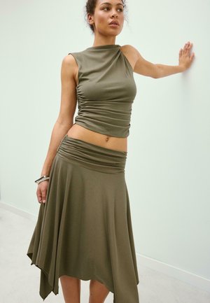 Žena sa opiera o bledú stenu, oblečená v bezrukávovom olivovo zelenom nariasenom topu a zodpovedajúcej asymetrickej splývavej sukni so striebornými náramkami.