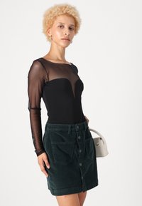 Svart langermet bodysuit i gjennomsiktig materiale med dyp V-hals, kombinert med en mørkegrønn kordfløyelskjørt med knapper foran og lommer.