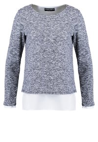 Grå stickad tröja med långa ärmar, rund hals och en något lös passform. Har ett strukturerat mönster och en lager-på-lager-look med ett vitt underlag.