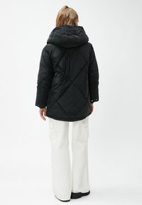 Person med lysebrunt hår i en hestehale iført en sort quilted hættejakke og hvide cargo bukser, stående mod en hvid baggrund.