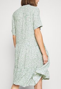 Abito floreale verde con motivi di margherite bianche, maniche corte, design a strati e orlo fluente leggermente arricciato.