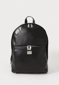 Zwarte leren rugzak met een gestructureerde vorm, voorzien van een voorvak, zilverkleurige ritsen en een metalen merkteken. Glad oppervlak.