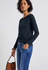 Frau mit lockigem Haar, die ein marineblaues Langarmshirt und blaue Jeans trägt, hält mit einer Hand eine braune Lederhandtasche und hat die andere Hand in der Tasche.